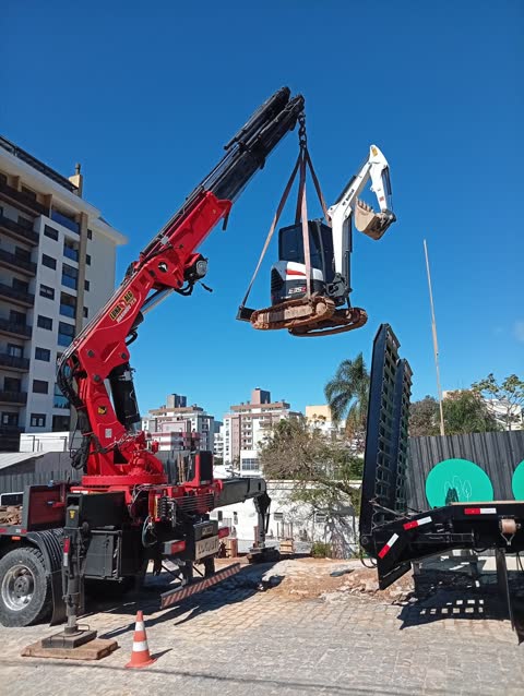 Guindaste articulado içando uma miniescavadeira em obra urbana
