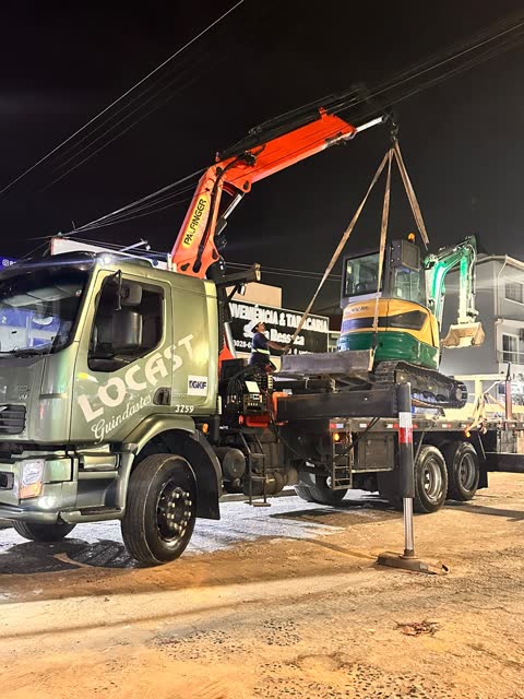 Caminhão da Locast com guindaste articulado içando uma escavadeira à noite
