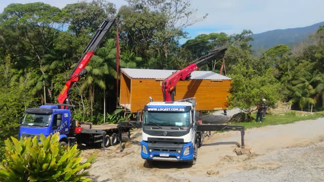 Dois guindastes articulados içando um módulo de madeira em área de mata