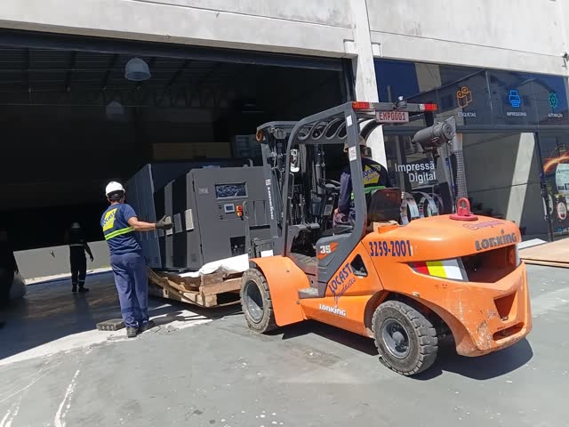 Empilhadeira levando máquina para dentro de galpão, com a equipe acompanhando